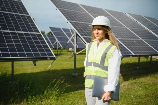 Портрет молодой женщины-инженера, стоящей возле солнечных панелей. Красивая женщина-профессионал в белом шлеме улыбается в камеру.
