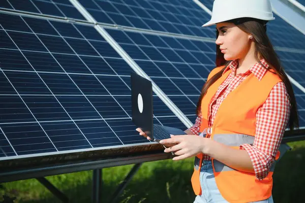 Portrait of electrician engineer in safety helmet and uniform using laptop checking solar panels. Female technician at solar station.
