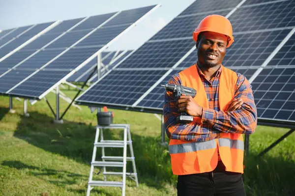 Gri tulumlu ve turuncu başlıklı ehil enerji mühendisi sahada yürürken güneş panellerini kontrol ediyor. Elinde enstrümanlarla pano ve konteynır taşıyan Afrikalı Amerikalı bir adam..