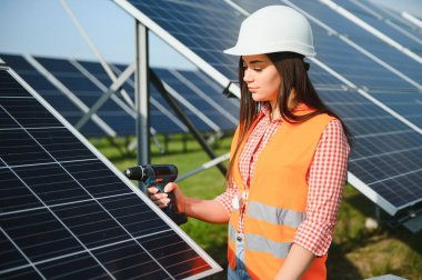 Portrait of electrician engineer in safety helmet and uniform using laptop checking solar panels. Female technician at solar station.