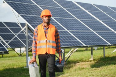 Koruyucu miğfer takmış yakışıklı Afrikalı Amerikalı zanaatkârın portresi. Üniformalı ve aletleri güneş panellerinin arasında duran bir adam..