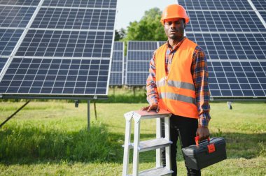 Koruyucu miğfer takmış yakışıklı Afrikalı Amerikalı zanaatkârın portresi. Üniformalı ve aletleri güneş panellerinin arasında duran bir adam..