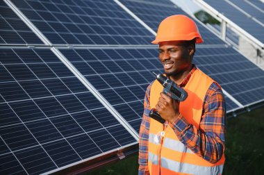 Güneş panellerinde tornavidayla güvenlik kaskı takan Afrikalı Amerikalı bir adam. İstasyonda servis çalışması yaparken araçları kullanan yetkili teknisyen.