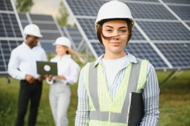 Çok ırklı insanlar ve güneş çiftliğine bakan ve çizimlere bakan güvenlik kaskları. İki mühendis ve teknisyen fotovoltaik inşaat planını açık havada inceliyor..