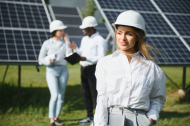 Çok ırklı insanlar ve güneş çiftliğine bakan ve çizimlere bakan güvenlik kaskları. İki mühendis ve teknisyen fotovoltaik inşaat planını açık havada inceliyor..