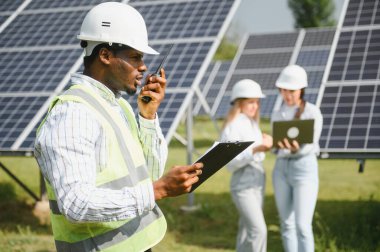 Çok ırklı insanlar ve güneş çiftliğine bakan ve çizimlere bakan güvenlik kaskları. İki mühendis ve teknisyen fotovoltaik inşaat planını açık havada inceliyor..