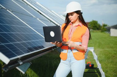 Portrait of electrician engineer in safety helmet and uniform using laptop checking solar panels. Female technician at solar station.
