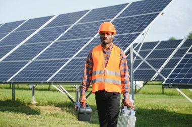 Gri tulumlu ve turuncu başlıklı ehil enerji mühendisi sahada yürürken güneş panellerini kontrol ediyor. Elinde enstrümanlarla pano ve konteynır taşıyan Afrikalı Amerikalı bir adam..