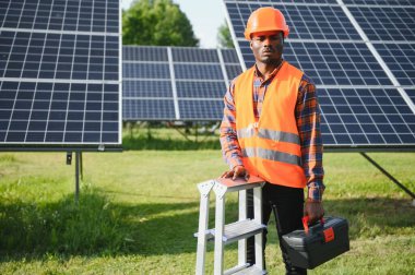 Gri tulumlu ve turuncu başlıklı ehil enerji mühendisi sahada yürürken güneş panellerini kontrol ediyor. Elinde enstrümanlarla pano ve konteynır taşıyan Afrikalı Amerikalı bir adam..
