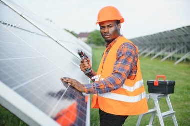 Gri tulumlu ve turuncu başlıklı ehil enerji mühendisi sahada yürürken güneş panellerini kontrol ediyor. Elinde enstrümanlarla pano ve konteynır taşıyan Afrikalı Amerikalı bir adam..