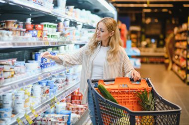 Marketteki raflar arasında alışveriş arabalarını iten genç bir kadın..