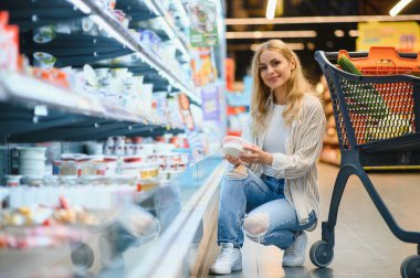 Genç havalı müşteri kadın süpermarket alışverişinde günlük kıyafet alışverişi yapıyor. Alışveriş arabasıyla alışveriş yapıyor. Hipermarketten ürünler seçiyor..