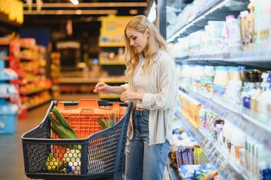 Genç havalı müşteri kadın süpermarket alışverişinde günlük kıyafet alışverişi yapıyor. Alışveriş arabasıyla alışveriş yapıyor. Hipermarketten ürünler seçiyor..