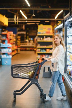 Marketteki raflar arasında alışveriş arabalarını iten genç bir kadın..