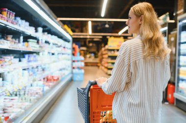 Genç havalı müşteri kadın süpermarket alışverişinde günlük kıyafet alışverişi yapıyor. Alışveriş arabasıyla alışveriş yapıyor. Hipermarketten ürünler seçiyor..