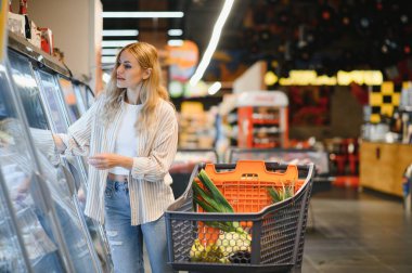 Genç havalı müşteri kadın süpermarket alışverişinde günlük kıyafet alışverişi yapıyor. Alışveriş arabasıyla alışveriş yapıyor. Hipermarketten ürünler seçiyor..