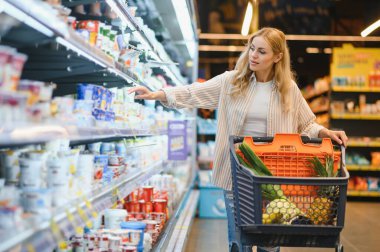 Genç havalı müşteri kadın süpermarket alışverişinde günlük kıyafet alışverişi yapıyor. Alışveriş arabasıyla alışveriş yapıyor. Hipermarketten ürünler seçiyor..