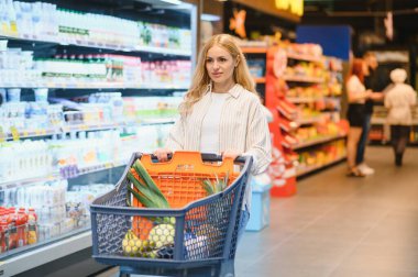 Marketteki raflar arasında alışveriş arabalarını iten genç bir kadın..