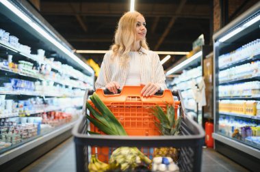 Marketteki raflar arasında alışveriş arabalarını iten genç bir kadın..