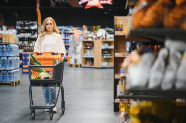 Gülümseyen mutlu genç kadın süpermarkette market arabasıyla günlük kıyafet alışverişinde..