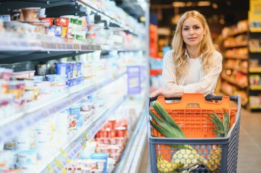 Marketteki raflar arasında alışveriş arabalarını iten genç bir kadın..