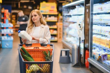 Genç havalı müşteri kadın süpermarket alışverişinde günlük kıyafet alışverişi yapıyor. Alışveriş arabasıyla alışveriş yapıyor. Hipermarketten ürünler seçiyor..