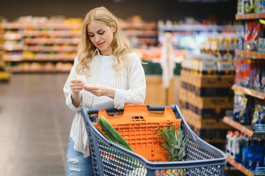 Marketteki raflar arasında alışveriş arabalarını iten genç bir kadın..