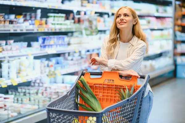 Marketteki raflar arasında alışveriş arabalarını iten genç bir kadın..