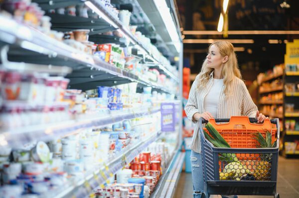 Молодая взрослая женщина толкает тележку между полками на рынке.