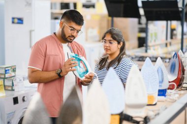 Young indian woman and a young man choosing a steam iron in a home appliances store. Cyber Monday concept. clipart