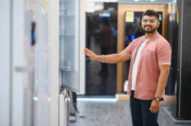 Smart modern indian male customer choosing large fridges in domestic appliances section. clipart