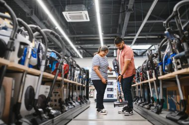 Teknoloji mağazasında elektrikli süpürgeyle rafın önünde çömelmiş genç bir çift..