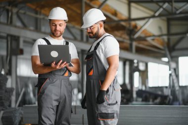 Fabrika işçisi ve mühendisi metal levha endüstriyel şantiyesinde birlikte çalışıyorlar, bilgisayardaki makine işlemini kontrol ediyorlar ve güvenlik için başlık takıyorlar.