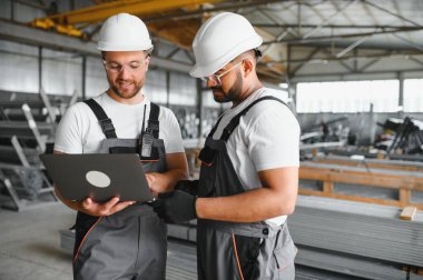 Fabrika işçisi ve mühendisi metal levha endüstriyel şantiyesinde birlikte çalışıyorlar, bilgisayardaki makine işlemini kontrol ediyorlar ve güvenlik için başlık takıyorlar.