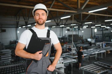Endüstriyel erkek mühendis, arkasında ağır bir fabrikada dururken beyaz kask takıyor. Endüstriyel makinelerde çalışmanın ve fabrikadaki güvenlik sistemini kontrol etmenin bakımı.