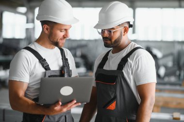 Fabrika işçisi ve mühendisi metal levha endüstriyel şantiyesinde birlikte çalışıyorlar, bilgisayardaki makine işlemini kontrol ediyorlar ve güvenlik için başlık takıyorlar.