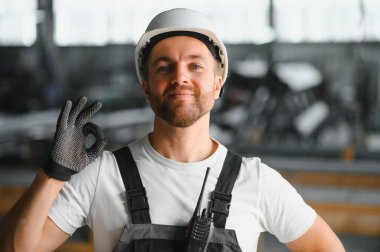 Endüstriyel erkek mühendis, arkasında ağır bir fabrikada dururken beyaz kask takıyor. Endüstriyel makinelerde çalışmanın ve fabrikadaki güvenlik sistemini kontrol etmenin bakımı.