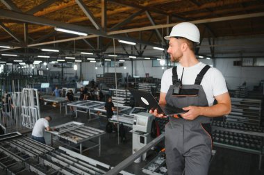 Endüstriyel erkek mühendis, arkasında ağır bir fabrikada dururken beyaz kask takıyor. Endüstriyel makinelerde çalışmanın ve fabrikadaki güvenlik sistemini kontrol etmenin bakımı.