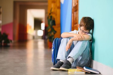 Zorbalık gördükten sonra yerde tek başına oturan küçük çocuk. Üzgün genç okul çocuğu koridorda elleri dizlerinin üstünde ve kafası bacaklarının arasında oturuyor..