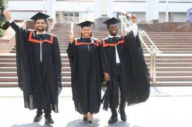 Mezuniyet törenine katılan çeşitli uluslararası öğrenciler mezuniyet şapkası ve cüppeli mutlu çok ırklı öğrenciler üniversite kampüsünde sıraya dizilmiş.