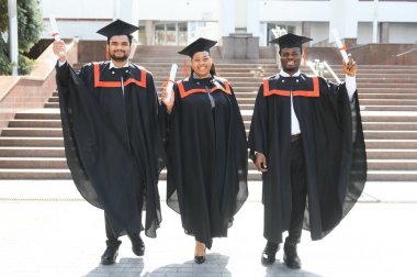 Mezuniyet elbiseleri ve şapkaları içinde, elinde diplomalarıyla mutlu, çok kültürlü bir grup insan kampüste kameraya bakarak üniversiteden mezun oluyor. Üniversite mezunu, üniversite konsepti.