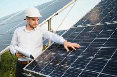 Kırsal kesimde güneşli bir günde beyaz gömlekli bir adam fotovoltaik panellerin yanında duruyor..