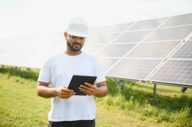 Genç Hintli erkek mühendisin portresi güneş panellerinin yanında duruyor, açık mavi gökyüzü arka planı, yenilenebilir ve temiz enerjisi var. beceri Hindistan, kopyalama alanı.