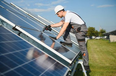 Erkek işçilerin güneş modülleri kurarken ve fotovoltaik güneş dizisini desteklerken yan görünümü. Güneş paneliyle çalışırken kaskını takan bir elektrikçi. Güneş enerjisi kavramı.