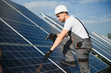 Elinde matkap, elinde tornavida olan üniformalı bir erkek işçi güneş panellerinin yanındaki bir merdiven üzerinde..