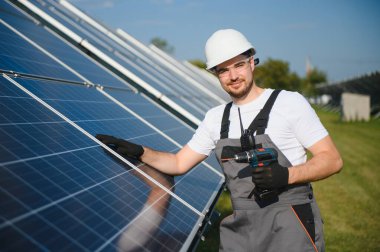 Erkek işçilerin güneş modülleri kurarken ve fotovoltaik güneş dizisini desteklerken yan görünümü. Güneş paneliyle çalışırken kaskını takan bir elektrikçi. Güneş enerjisi kavramı.