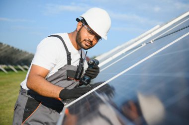 Mavi gökyüzüne karşı güneş panellerinin yanında duran beyaz şapkalı genç Hintli teknisyen portresi. Endüstriyel işçi güneş sistemi kurulumu, yenilenebilir yeşil enerji üretimi kavramı