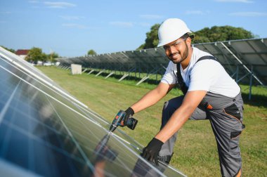 Mavi gökyüzüne karşı güneş panellerinin yanında duran beyaz şapkalı genç Hintli teknisyen portresi. Endüstriyel işçi güneş sistemi kurulumu, yenilenebilir yeşil enerji üretimi kavramı