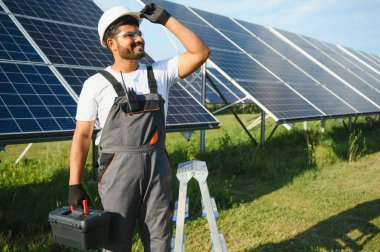 Mavi gökyüzüne karşı güneş panellerinin yanında duran beyaz şapkalı genç Hintli teknisyen portresi. Endüstriyel işçi güneş sistemi kurulumu, yenilenebilir yeşil enerji üretimi kavramı