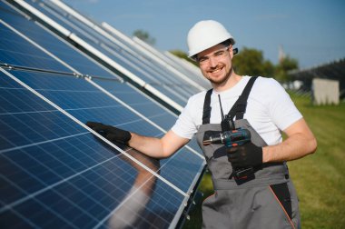 Male worker in uniform with a drill - screwdriver in his hand on a stepladder near solar panels. clipart